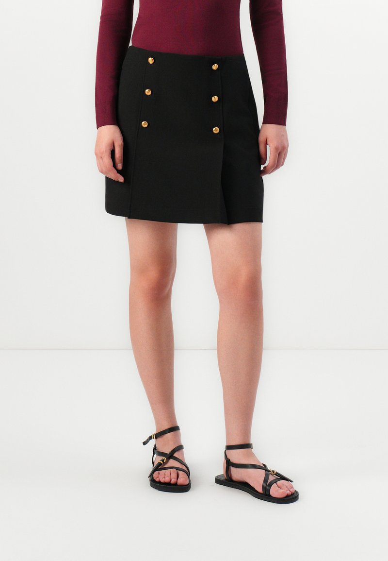 Jupe mini noire avec des accents de boutons dorés, conçue avec un devant croisé et une coupe droite. Associée à des sandales noires à lanières.