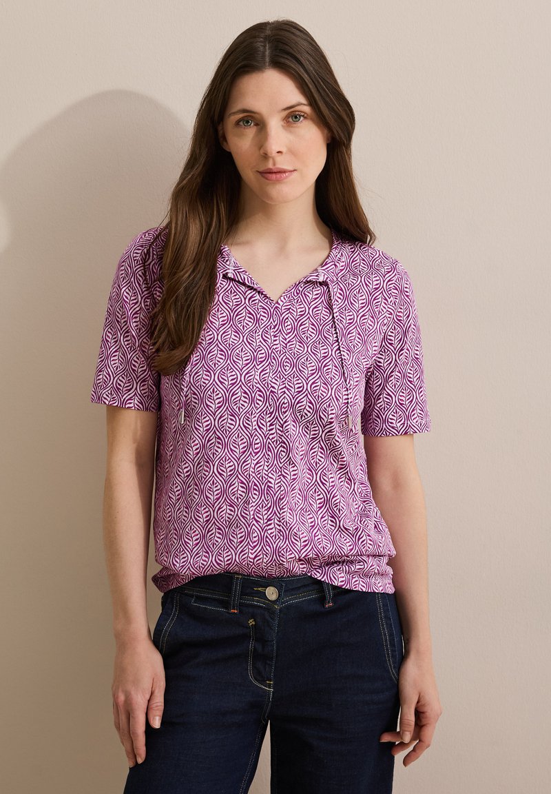 Blusa morada de manga corta con un estampado de hojas, que cuenta con un cuello con muesca y una tela suave. Combinada con pantalones de mezclilla azul oscuro.