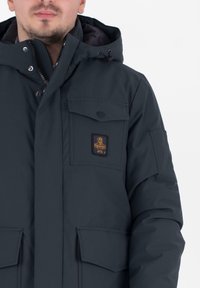 Giacca blu scuro con cappuccio, dotata di zip frontale, molteplici tasche e un patch con logo sul petto. Realizzata in tessuto liscio e resistente all'acqua.
