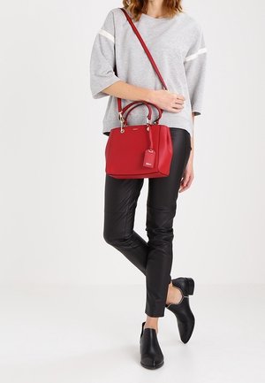 Femme portant un haut gris et un pantalon noir, tenant un sac à main rouge avec des poignées supérieures et une bandoulière, debout sur une jambe.