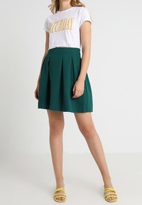 Bílý tričko s nápisem "SOBOTA" v žlutém tisku, v kombinaci s průsvitnou sukní do kolen v teal barvě a žlutými sandály na řemínky.