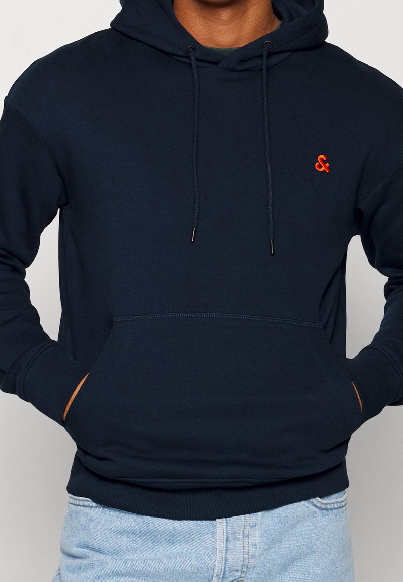 Marinblå bomullshuva med en känguruficka, justerbara dragkedjor och en liten orange logotyp på bröstet. Mjuk textur.