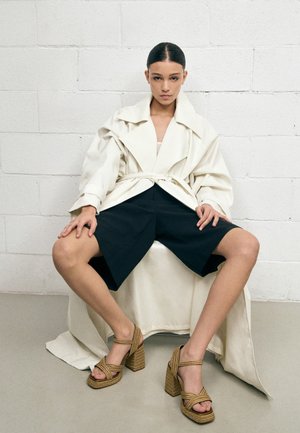 Kvinne med hår greid bakover, iført oversized hvit jakke, svarte knelang shorts og lysebrune høye hæler, sittende mot en hvit murvegg.