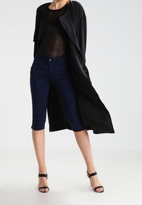Musta pitkä takki, läpinäkyvä musta verkkopaita, tummansiniset capri-housut ja mustat korkokengät. Sleek-materiaalit ja istuva muotoilu.