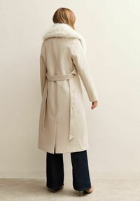 Abrigo largo beige con un cuello de piel sintética, cintura ceñida, aberturas laterales y una textura suave, combinado con jeans oscuros de pierna acampanada y tacones.