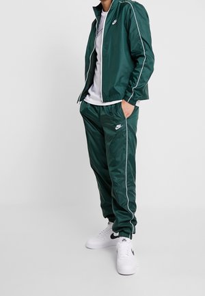 Osoba ubrana w ciemnozielony dres Nike z białymi lamówkami, pod którą ma białą koszulkę, oraz w białe buty Nike, stoi z jedną ręką w kieszeni.