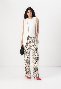 Light beige vest with buttons over white top, paired with cream trousers featuring black floral patterns and red high-heeled shoes. Black bag.