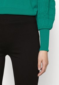 Turquoise gebreide trui met gestructureerde poffende mouwen, gecombineerd met zwarte leggings. De trui heeft geribde manchetten en een gladde afwerking.