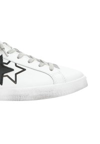 Sneaker bianco con lacci grigi e un patch a forma di stella nera sul lato, caratterizzato da una suola bianca leggermente usurata.