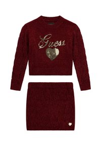 Ensemble pull en maille rouge et jupe assortie, avec un motif en maille torsadée, un logo "Guess" doré en sequins, et un accent en forme de cœur sur la jupe.