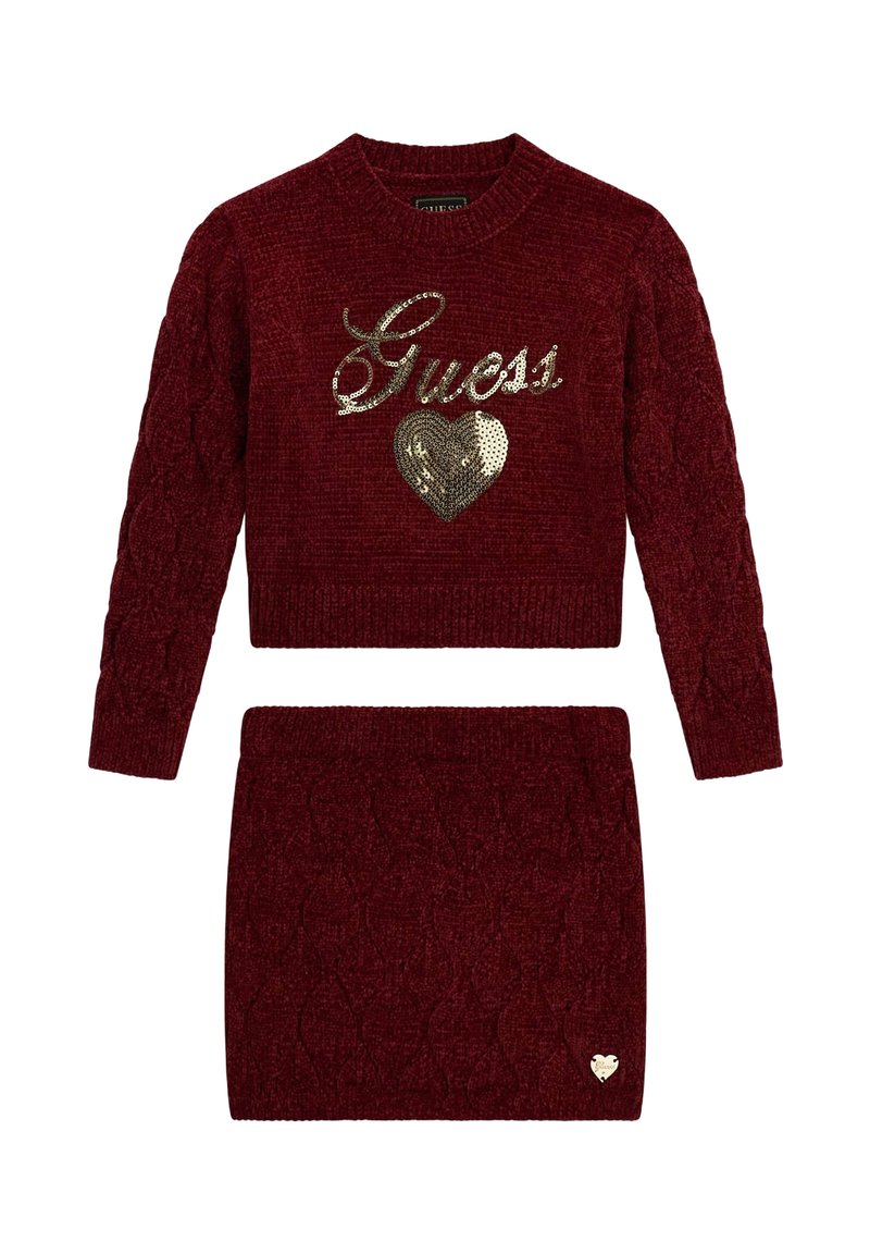 Ensemble pull en maille rouge et jupe assortie, avec un motif en maille torsadée, un logo "Guess" doré en sequins, et un accent en forme de cœur sur la jupe.