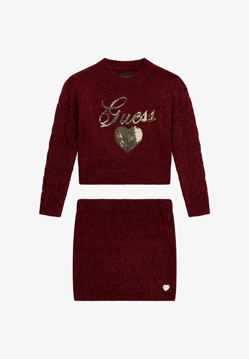 Ensemble pull en maille rouge et jupe assortie, avec un motif en maille torsadée, un logo "Guess" doré en sequins, et un accent en forme de cœur sur la jupe.