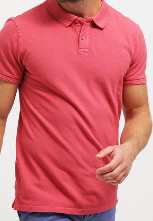 Muž v přiléhavé korálově zbarvené polokošili s krátkými rukávy a zapnutým límečkem, modrých kalhotách, uprostřed akce s pravou rukou zvednutou.