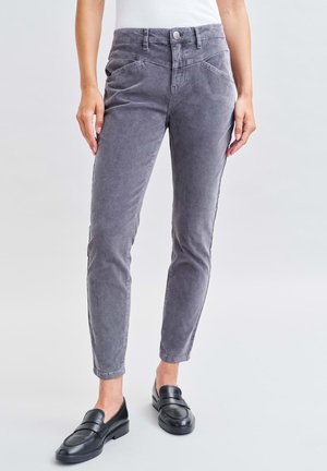 Vrouw draagt strakke grijze broek en zwarte loafers, staand tegen een effen lichte achtergrond.