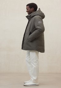 Giacca imbottita verde oliva con cappuccio, chiusura con cerniera, tasche laterali e logo sulla manica sinistra. Indossata con pantaloni bianchi e scarpe da ginnastica.