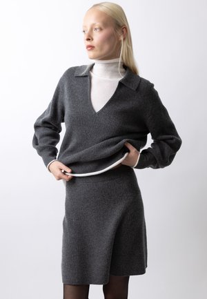 Ung kvinde iført en mørkegrå ribstrikket sweater og matchende nederdel, justerer sweaterens kant over en lys rullekrave.