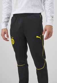 Man in zwarte sportbroek met een gele Puma-logo en het embleem van Borussia Dortmund, gecombineerd met een wit langesleeve shirt.