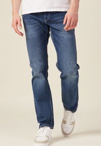 Blaue Jeans im geraden Schnitt mit ausgewaschenen Details und kontrastierenden Nähten, kombiniert mit weißen und beigen Sneakern.