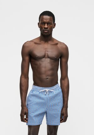 Άνδρας χωρίς πουκάμισο στέκεται μπροστά σε απλό φόντο, φοράει μπλε και λευκά καρό μαγιό με λευκό κορδόνι.