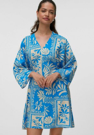 Donna che indossa un vestito fantasia floreale blu e bianco al ginocchio con maniche lunghe e ampie, in piedi con le mani incrociate in vita.