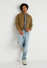 Καφέ σακάκι πάνω από γκρι μπλούζα, συνδυασμένο με ανοιχτό γαλάζιο τζιν και μπεζ αθλητικά παπούτσια. Το μοντέλο στέκεται μπροστά σε λευκό φόντο.