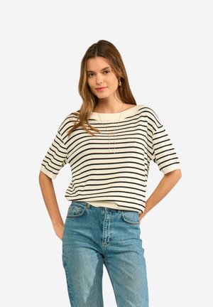 Jonge vrouw met een wit kortemouwtop met zwarte horizontale strepen en blauwe high-waisted jeans, staat met handen in de zij.