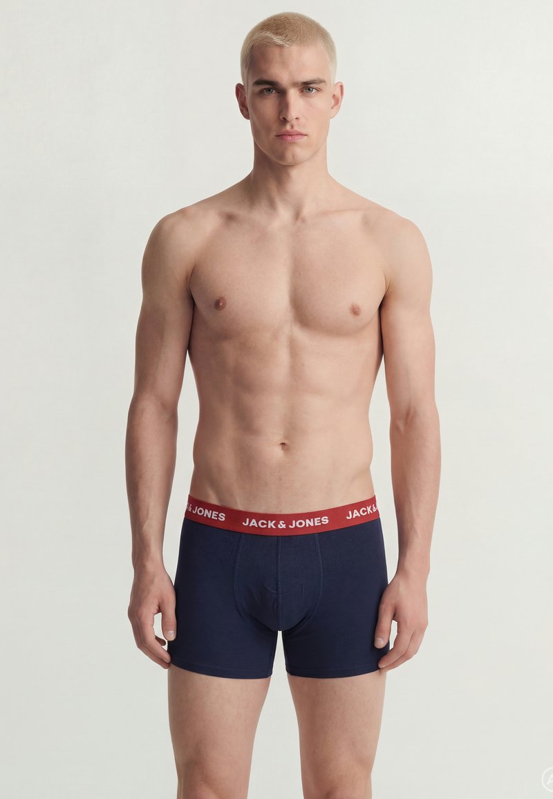 Jeune homme aux cheveux blonds courts se tient de face, portant un boxer bleu marine avec une ceinture rouge portant l'inscription "JACK & JONES" sur un fond uni.