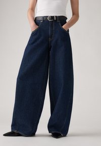 Wijde blauwe denim jeans met een hoge taille, twee voorzakken, gouden stiksels en een zwarte riem met een zilveren gesp. Zwarte puntige schoenen zichtbaar.
