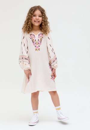 Lächelndes Mädchen mit lockigem Haar, das ein weißes Kleid mit bunter Blumenstickerei und weiße Turnschuhe trägt und vor einem einfarbigen Hintergrund steht.