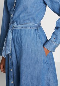 Vaaleansininen farkkumekko, jossa pitkät hihat, napitettu edusta, vyötärön nyöri ja sivutaskut. Hihansuissa näkyy brändin tunnus.