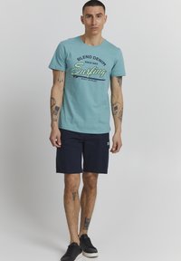 Turkuza krāsas kokvilnas t-krekls ar tumši zaļu "Blend Denim Surfing" grafiku, apvienots ar tumši zilām šortiem un melniem sporta apaviem.