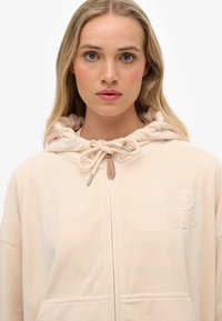 Haut à capuche beige doux avec cordon de serrage, fermeture éclair frontale et détail de logo en relief sur la poitrine. Design uni avec poches.