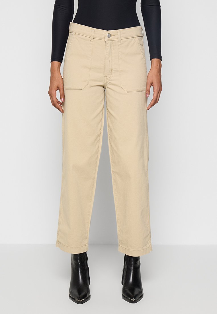 Pantalones beige de pierna ancha hechos de algodón, con bolsillos frontales, cierre de botón y un ajuste holgado, combinados con botas de tacón negras.