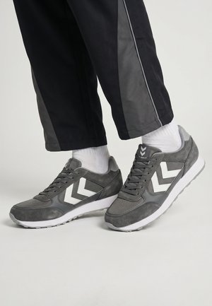 Person trägt grau-weiße Sneakers mit Chevron-Logo, weiße Socken und schwarze Hosen mit grauen Seitenstreifen, steht auf hellem Boden.