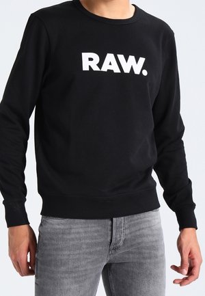 Osoba ubrana w czarną bluzę z długim rękawem z białym napisem „RAW.” na piersi i jasnoszare dżinsy.