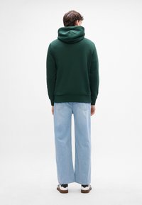 Felpa verde scuro con cappuccio regolabile e polsini a coste, abbinata a jeans a gamba larga blu chiaro. Sneakers bianche visibili nella parte inferiore.