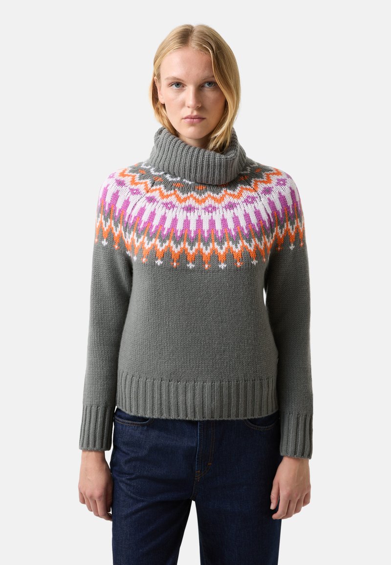 Žena v šedém roláku s geometiickým vzorem v růžové, oranžové, bílé a fialové barvě a tmavě modrých džínách stojící na jednobarevném pozadí.