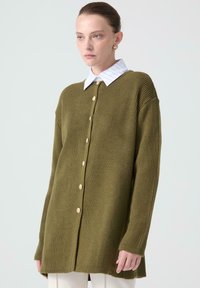 Cardigan lavorato a maglia in verde oliva con texture a coste, dotato di una camicia con colletto sotto, bottoni frontali e una vestibilità ampia.