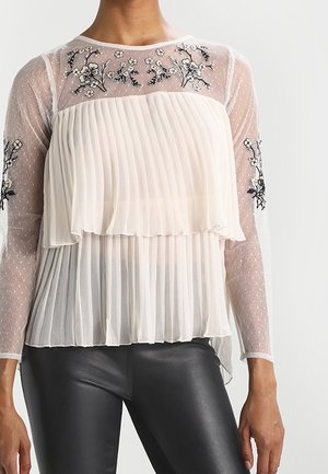 Hvid transparent plisseret bluse med blomsterbroderi på bryst og ærmer, båret sammen med sorte læderbukser.