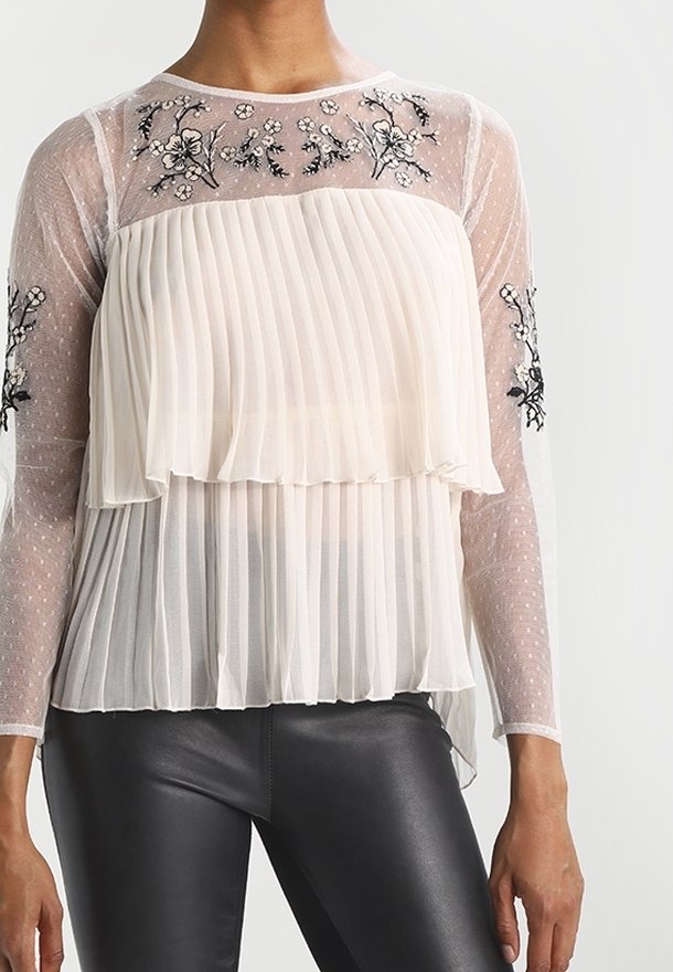Blouse blanche plissée et transparente avec broderie florale sur le buste et les manches, portée avec un pantalon en cuir noir.