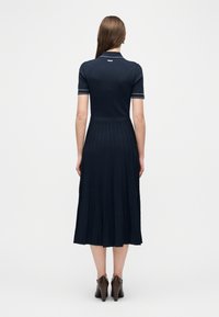 Γυναίκα που στέκεται με την πλάτη στραμμένη, φορώντας ένα κοντομάνικο φόρεμα σε ναυτικό μπλε με πλισέ φούστα και καφέ παπούτσια με τακούνι, σε λευκό φόντο.