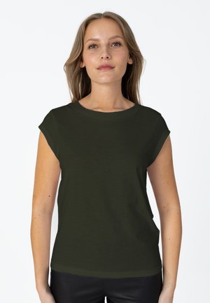 Jeune femme aux longs cheveux portant un t-shirt uni vert foncé à manches courtes et un pantalon noir, debout devant un fond blanc.