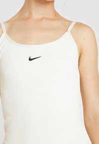 Canotta bianca a coste con spalline sottili, caratterizzata da un piccolo logo Nike nero al centro. Tessuto liscio con un design aderente.