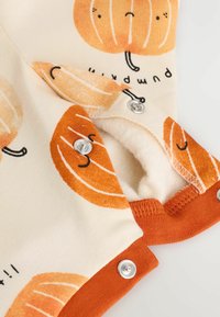 Tissu imprimé de citrouille en crème avec des accents orange ; présente des fermetures à pression et une doublure intérieure douce. Le texte comprend "citrouille".