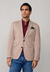 Helle beige Blazer mit zwei Knöpfen, Revers und Fronttaschen, getragen über einem dunkelroten Polo und blauen Jeans, mit einem gemusterten Einstecktuch.