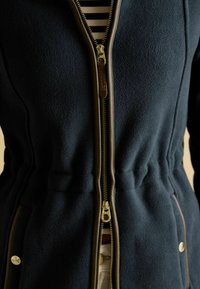 Námornícky fleecový bundá s kovovým zipsom, sťahovaním v páse a béžovými akcentmi na bokoch. Texturovaná látka a detaily zlatého tónu.