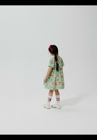 Ung pige med langt fletningshår iført en blomsterkjole, hvide sneakers, stribede sokker og et rødt pandebånd, stående på en ensfarvet hvid baggrund.