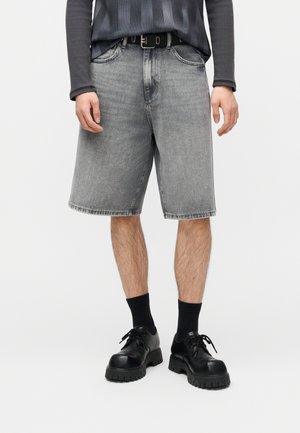 Persona che indossa pantaloncini di denim grigio sbiadito, cintura nera, calzini neri e scarpe nere robuste con lacci su uno sfondo bianco.