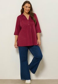 Camicia bordeaux con maniche a tre quarti, caratterizzata da pieghe, indossata con jeans blu a gamba larga e sneakers bianche. Tessuto dalla texture liscia.