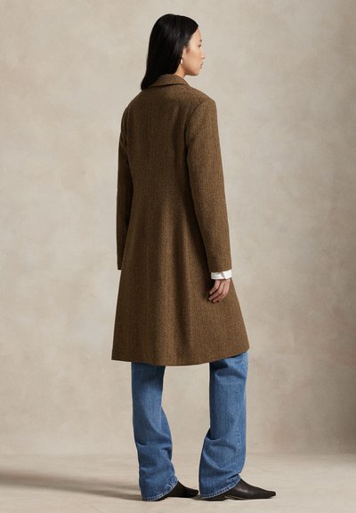 Kobieta stojąca zwrócona w prawo, ubrana w długi brązowy płaszcz w jodełkę, niebieskie dżinsy i czarne, spiczaste buty na neutralnym tle.